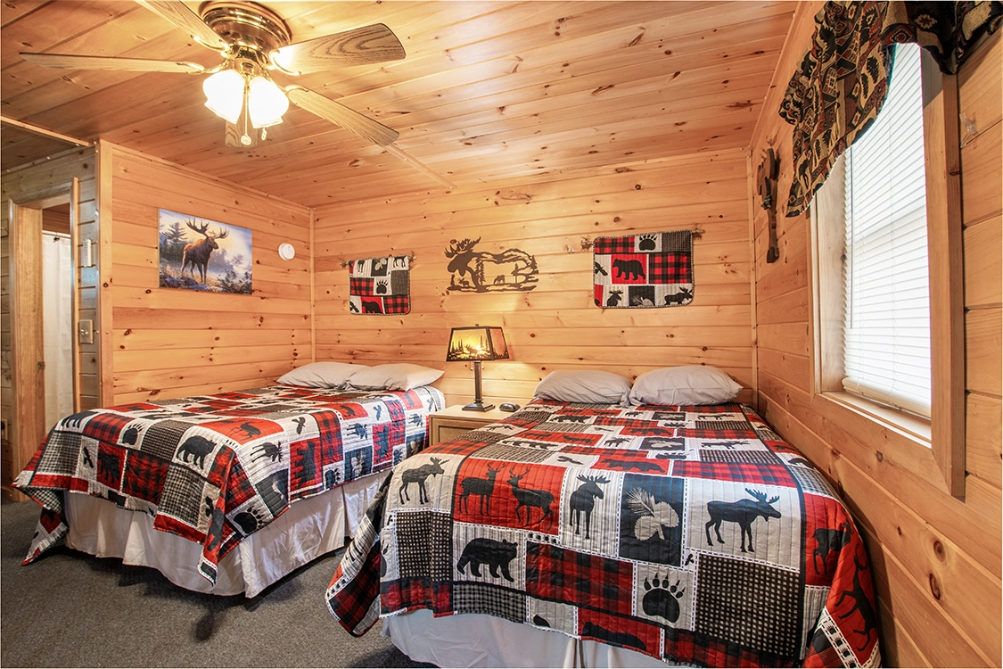 Cozy cabin bedroom with wood-paneled walls, two beds with patchwork quilts featuring moose and bear designs, a nightstand with a lamp, ceiling fan, window with patterned valance, and rustic wildlife decor.